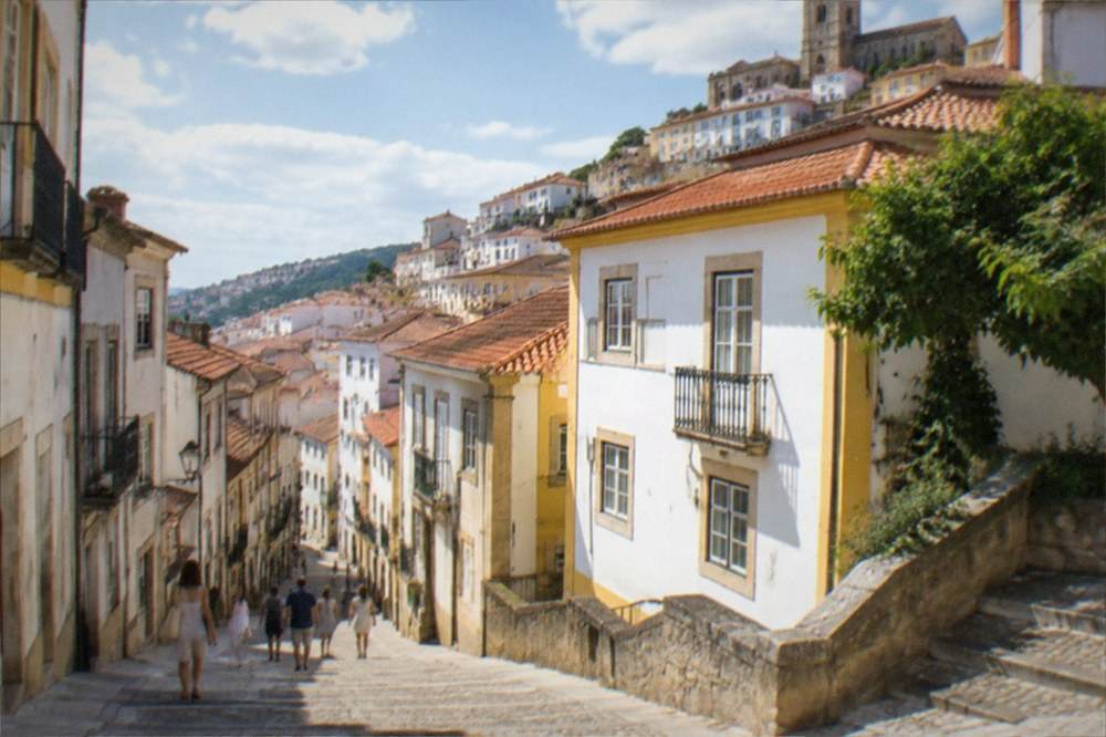 Visitors in light clothing walking along a sunlit street in the old town of Coimbra in warm weather