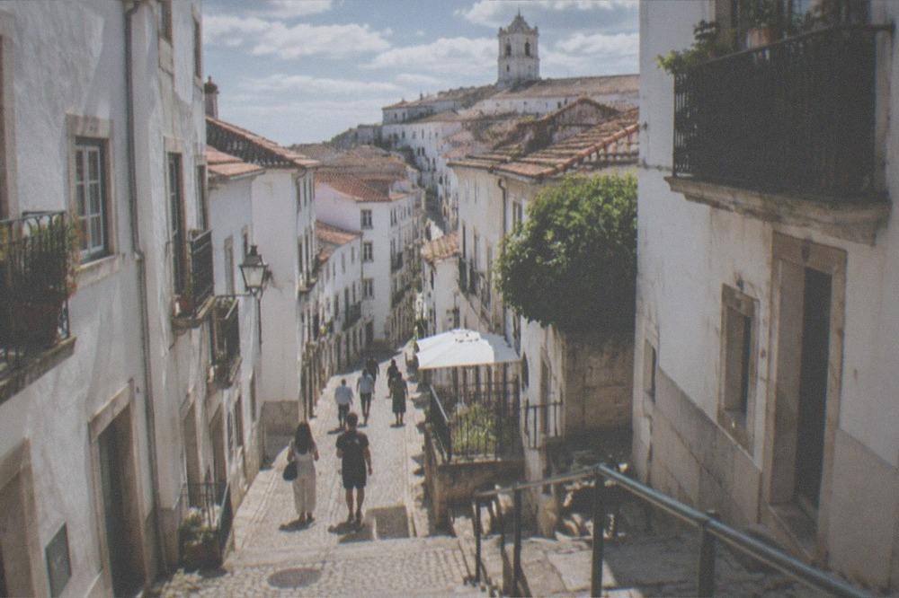 Visitors walking along a sunlit cobbled street in the upper town of Coimbra in warm weather.