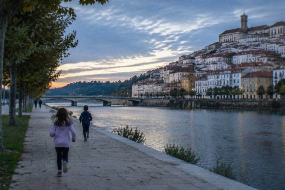 Children running on a wide flat riverside path with trees and the city bridge in the background